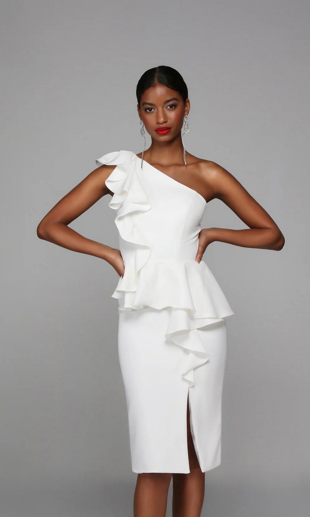 A woman stands against a plain grey background wearing the Alyce 70007: a knee-length white party dress featuring an elegant one-shoulder ruffle sheath design with a front slit.